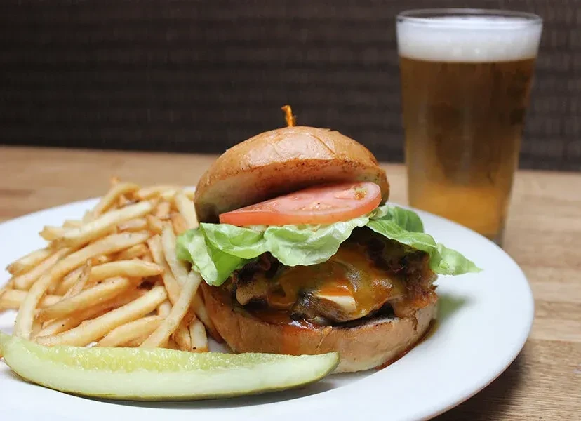 Cheeseburger, fries, pickle, and beer on a wooden table.
