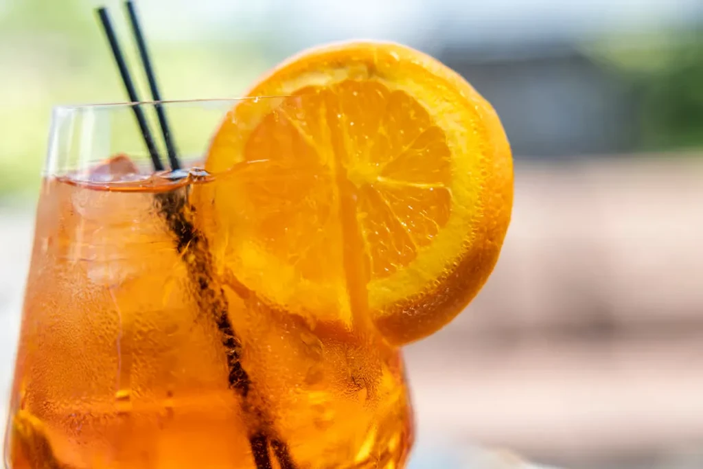 Orange cocktail in clear glass with ice, orange slice, and straws.