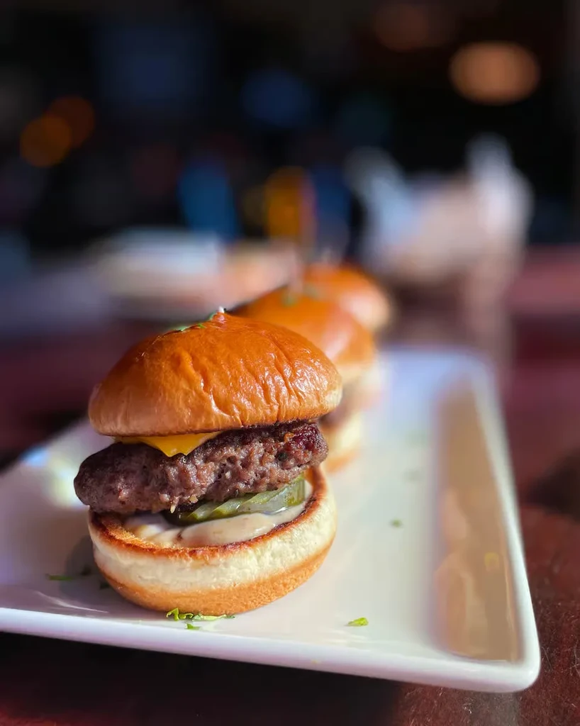 Line of mini-burgers with beef, melted cheese, pickles, and sauce.