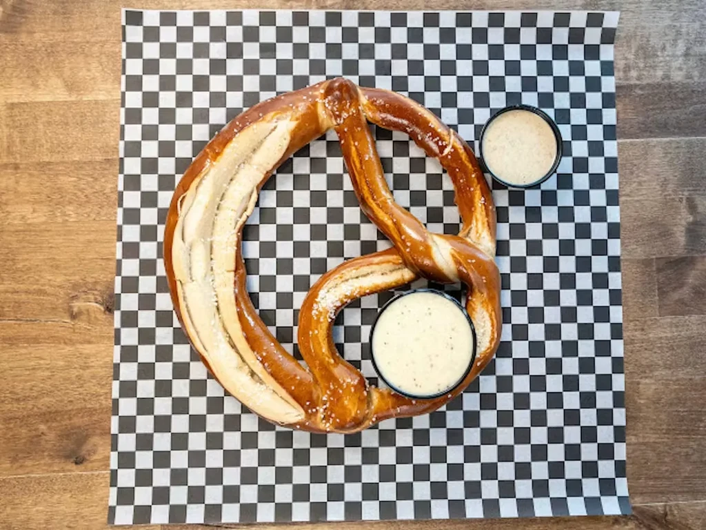 Golden-brown soft pretzel, salted, with two dipping sauces on checkered paper.
