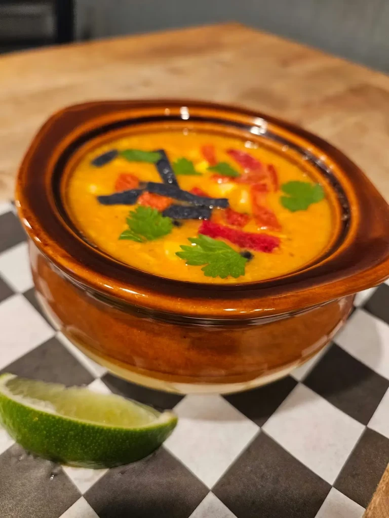 Close-up of orange soup with tortilla strips, peppers, cilantro, and lime.
