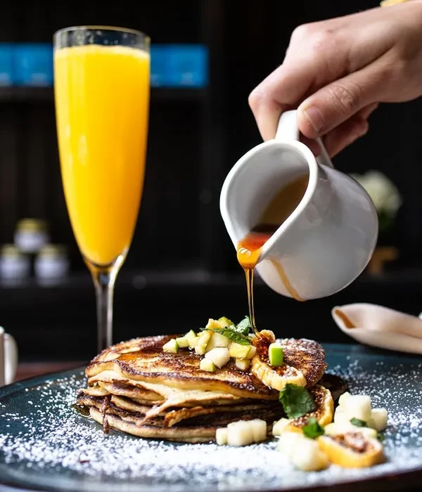 Hand pours syrup over pancakes with fruit, mint, and orange drink.