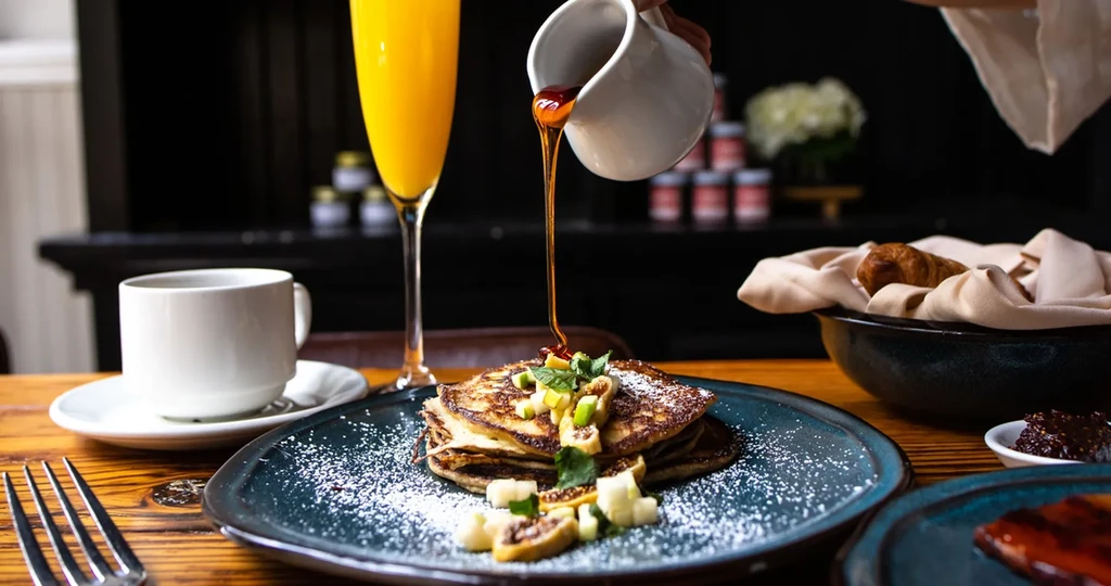 Pancakes with fruit, pouring syrup, mimosa on rustic wooden table.