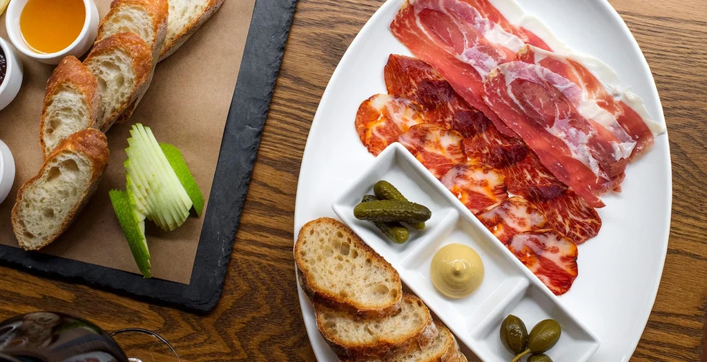 Charcuterie board with cured meats, baguette, pickles, and apple on wood.