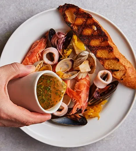 Hand pouring herbed sauce onto a generous seafood platter.