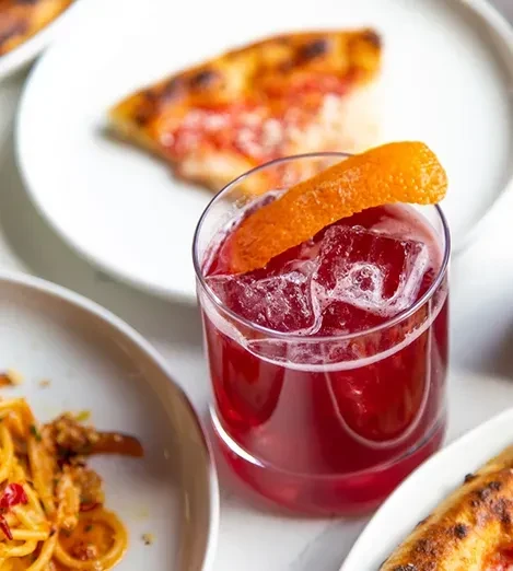 A red cocktail with orange peel, surrounded by pizza and pasta.