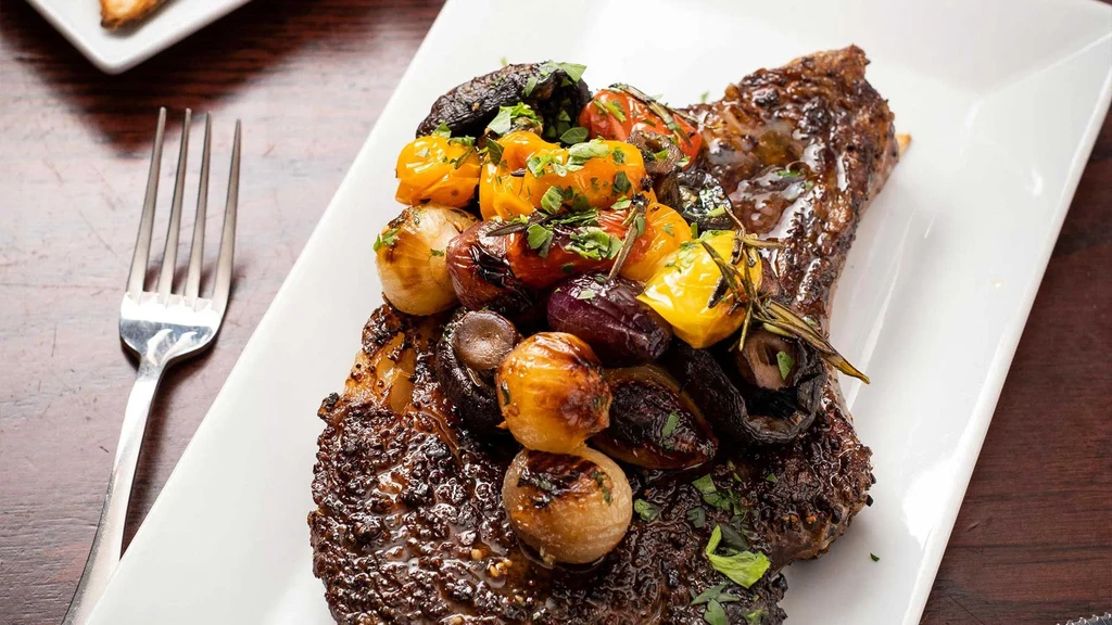 Seared steak topped with roasted vegetables and herbs on a white plate.