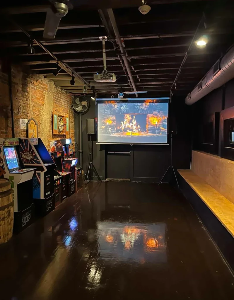 Dimly lit industrial game room with arcade games and projected fireplace.