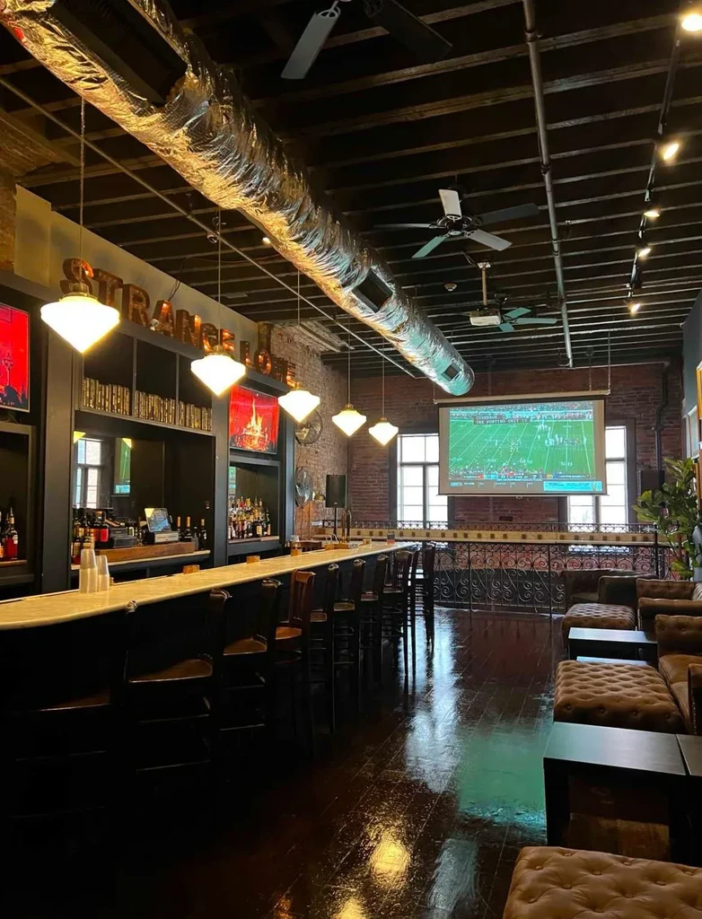 Modern industrial bar with 'STRANGE LOVE' sign, leather sofas, and sports screen.