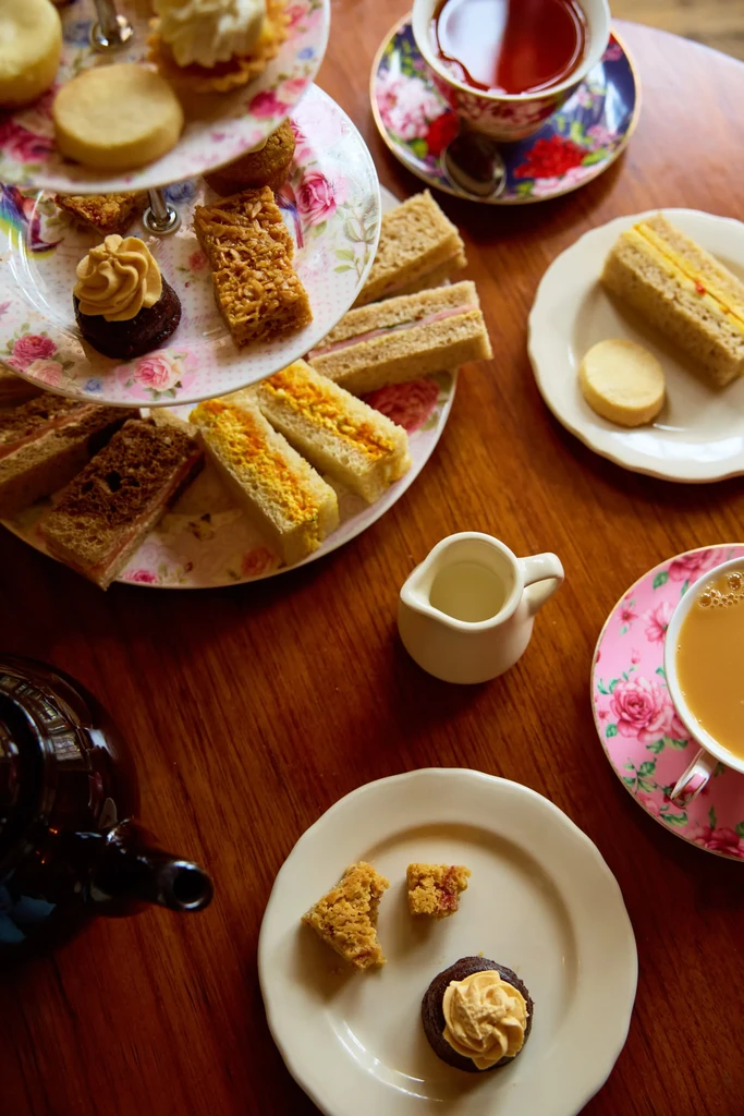 Tiered stand of afternoon tea pastries, sandwiches, and teacups on a table.