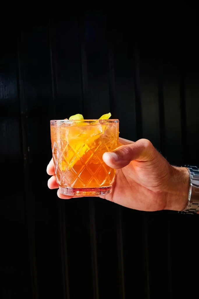 Hand holds amber cocktail with ice and citrus in a faceted glass.