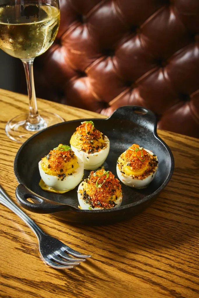 Four deviled eggs with roe and spices in a skillet, beside white wine.