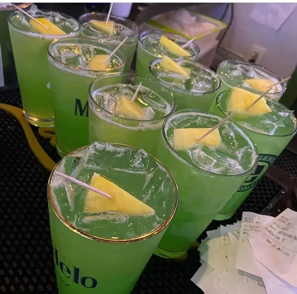 Nine branded green drinks with ice and fruit on a bar.