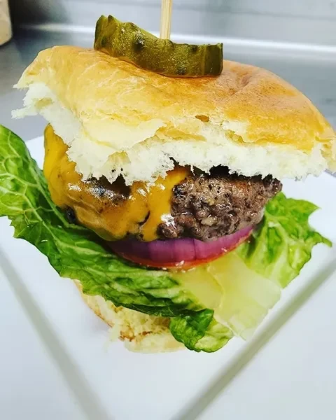 Cheeseburger with melted cheese, lettuce, onion, pickle spear, on white plate.