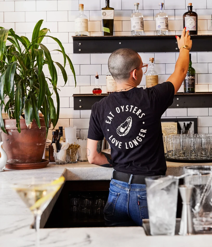 Bartender in oyster shirt reaches for bottles; martini glass on counter.