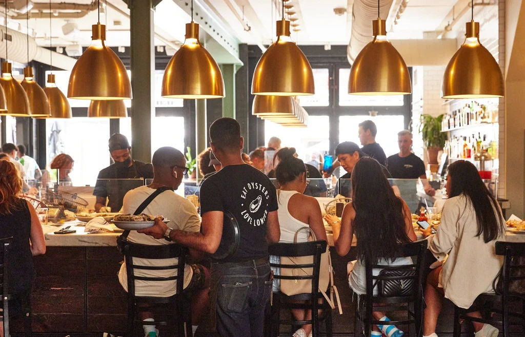 Modern restaurant bar interior: patrons, server, brass pendant lights.