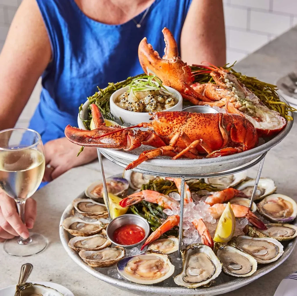 Two-tier seafood tower with cooked lobster, oysters, shrimp, and wine.