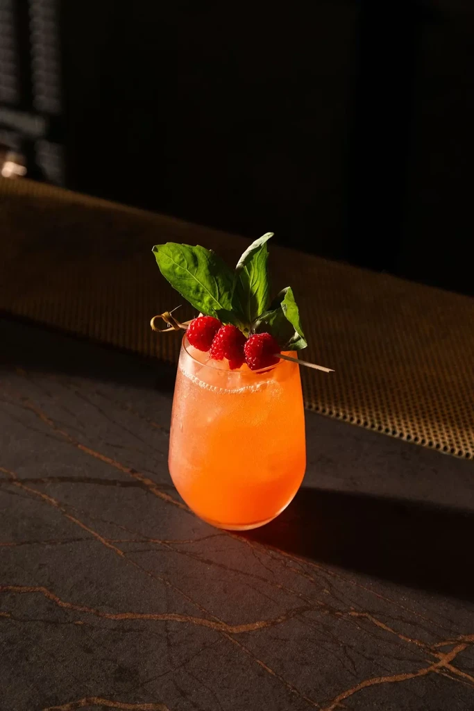 Orange cocktail in stemless glass with raspberries, green leaves, on dark marble.