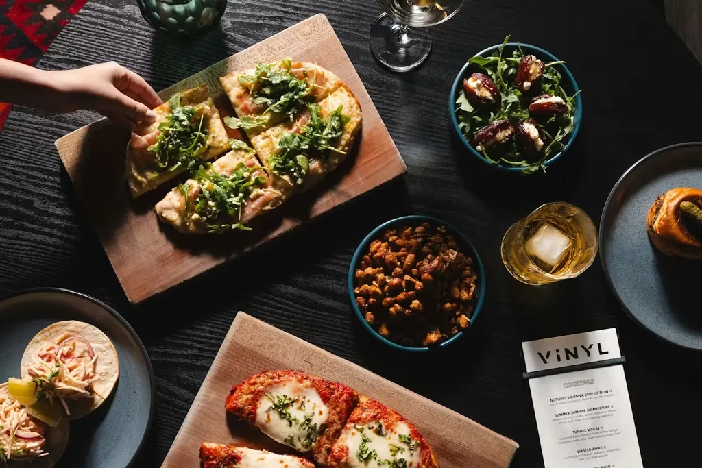 Overhead shot of dark table with flatbread, tacos, stuffed dates, and drinks.
