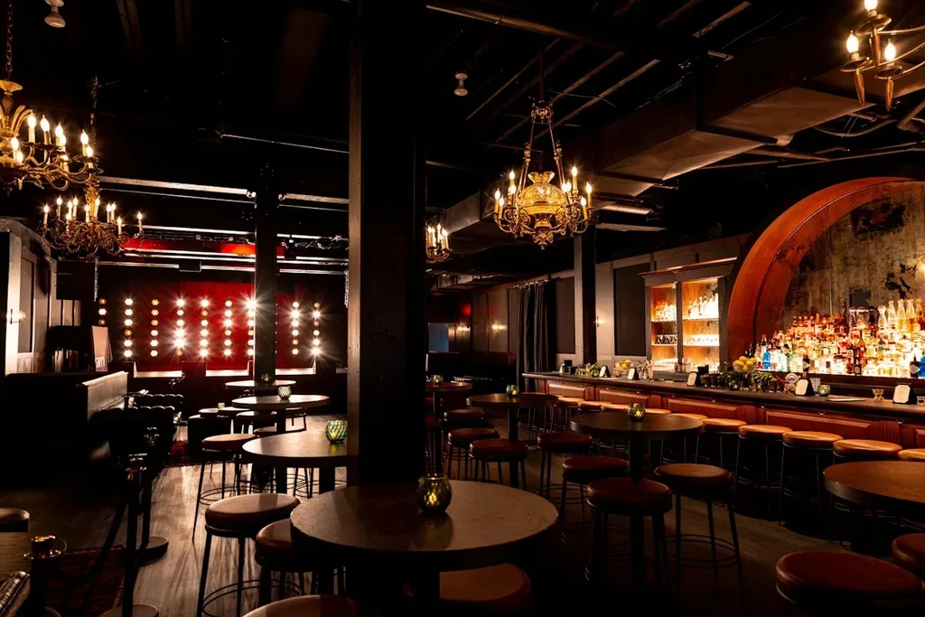 Dimly lit bar interior with tables, stocked bar, and ornate chandeliers.