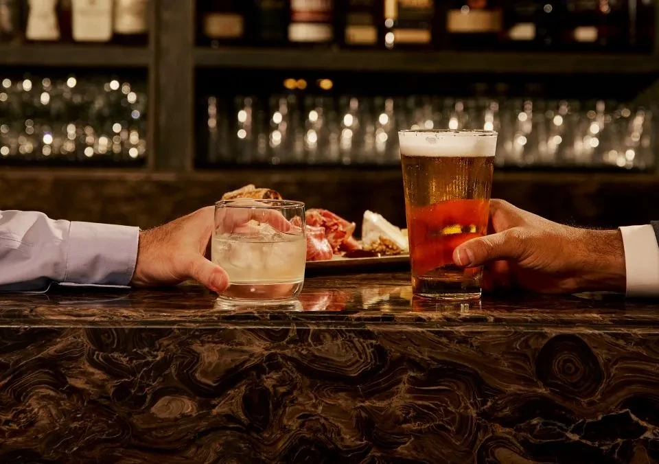 Hands hold cocktail, beer, with charcuterie on a polished bar.