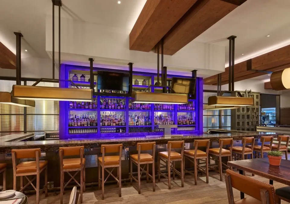 Empty modern bar, purple-lit alcohol display, high stools, wood beams.