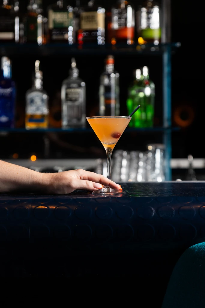 Orange cocktail in martini glass with red garnishes and a hand.