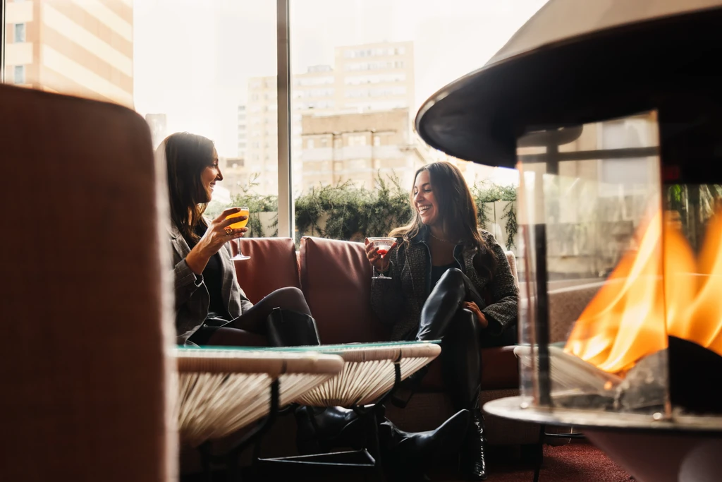 Two women laugh over drinks in an upscale lounge with fireplace.