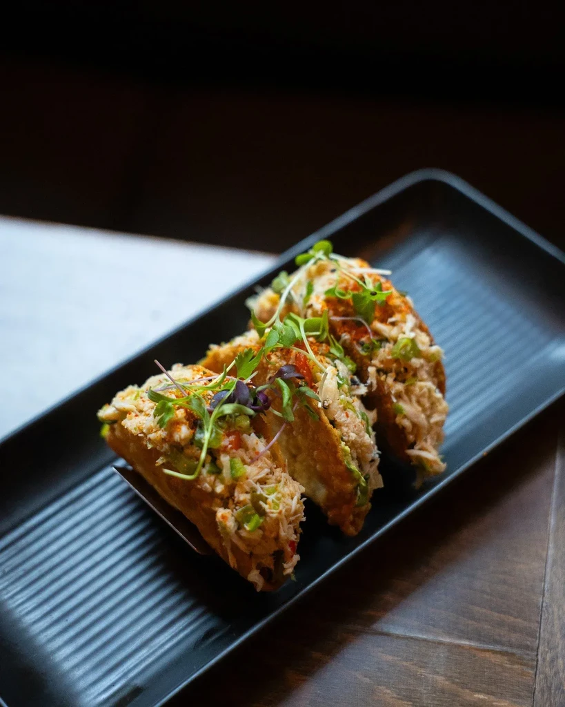 Three crispy taquitos with microgreens on a black rectangular plate.