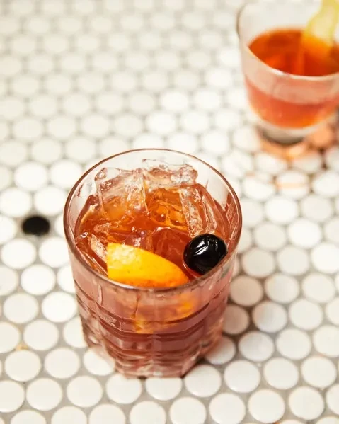 Amber cocktail with ice, orange peel, and cherry on a tiled surface.