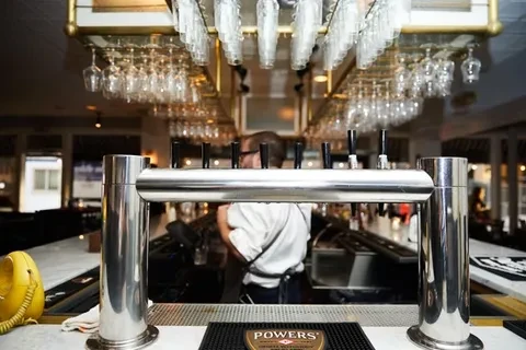 Bar counter with stainless steel taps, hanging glassware, and a bartender.
