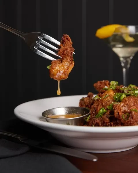 Fork holds sauced fried chicken dripping over a plate and martini.