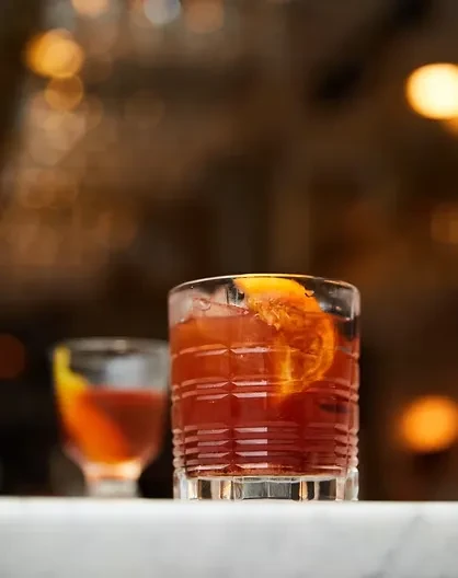 Reddish-orange drink in clear textured glass with orange slice on bar.
