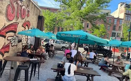 Lively outdoor patio with picnic tables, teal umbrellas, and people.