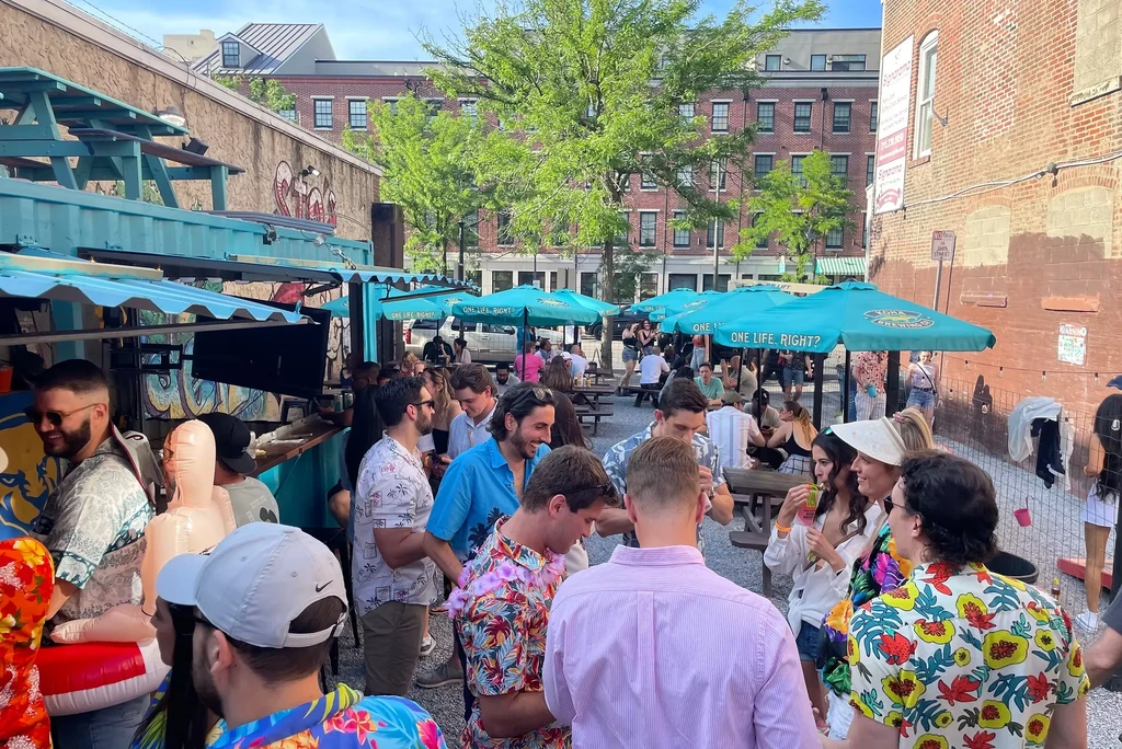 Bustling outdoor bar patio with people, turquoise container bars, and umbrellas.