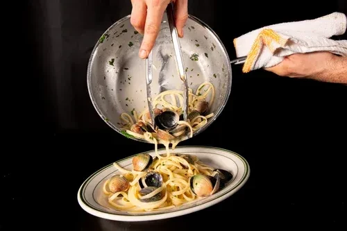 Hands serving pasta with clams from a pan onto a plate.