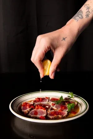 Hand squeezes lemon over sliced seared tuna with red sauce.