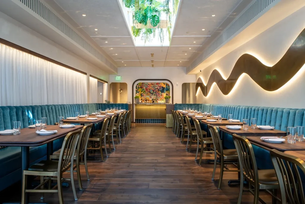 Empty modern restaurant dining room with tables, teal booths, plants, bar.