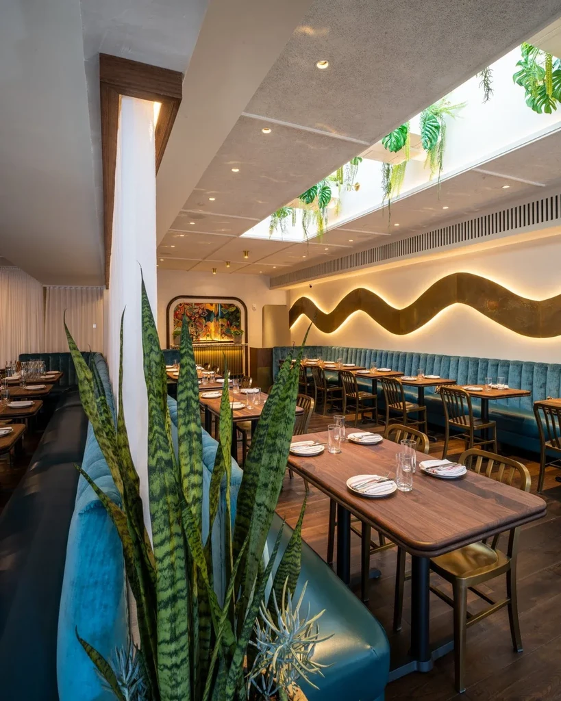 Modern restaurant interior with dark tables, teal banquettes, and plants.