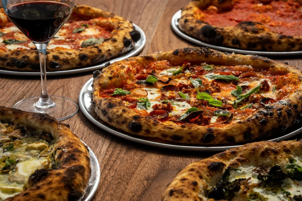 Five distinct Neapolitan pizzas on a wooden table with red wine.