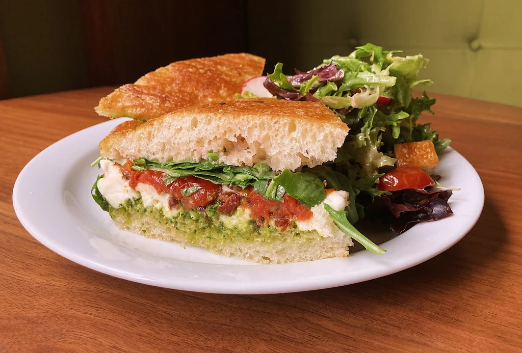 Rustic focaccia sandwich with pesto, mozzarella, tomatoes, arugula, and a side salad.