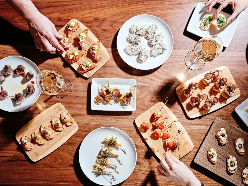 Hands reaching for gourmet appetizers and white wine on a wooden table.