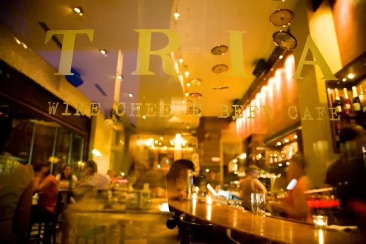TRIA WINE CHEESE & BEER CAFE on glass, with warm, blurry cafe interior.
