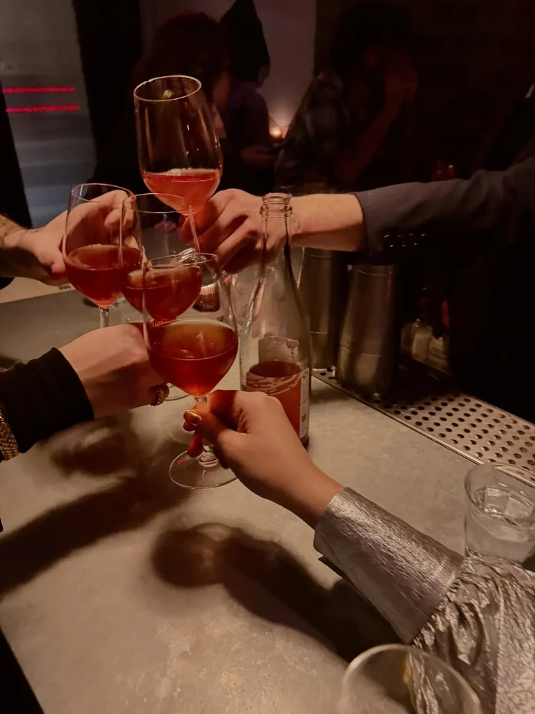Four hands toast with drinks in wine glasses at a dimly lit bar.