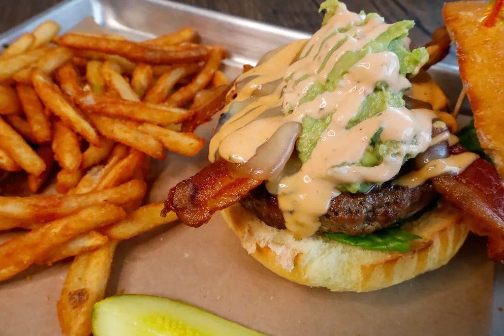 Gourmet burger with bacon, guacamole, fries, and a pickle spear.