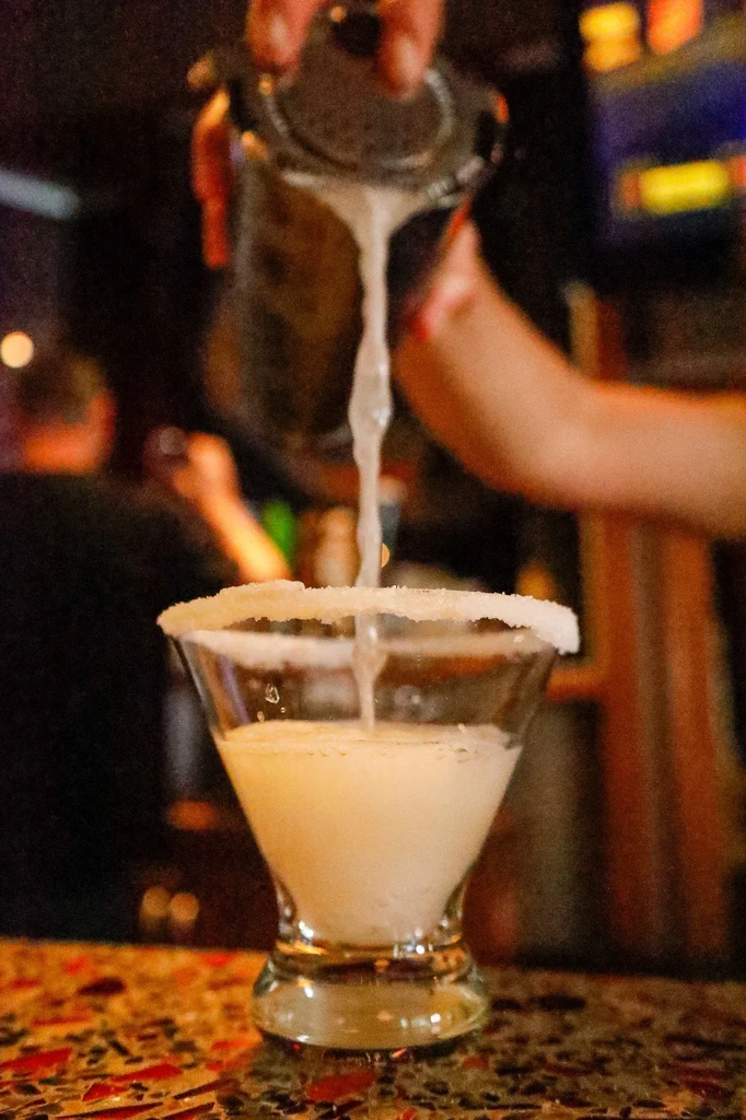 Hands pour frothy cocktail from shaker into a salt-rimmed martini glass.
