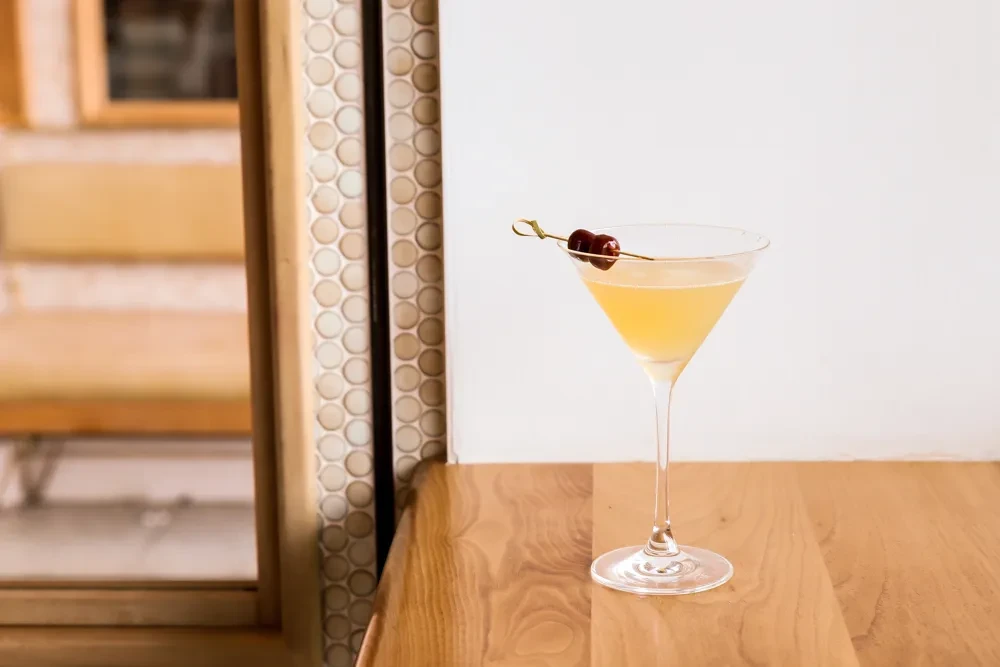 Light amber cocktail in martini glass, garnished with two cherries.