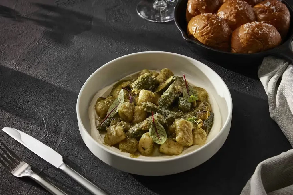 Gnocchi with green sauce, dinner rolls, and cutlery on a dark table.