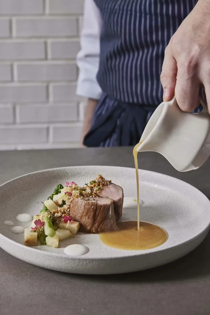 Professionally plated sliced meat, vegetables, flowers, and pouring sauce.