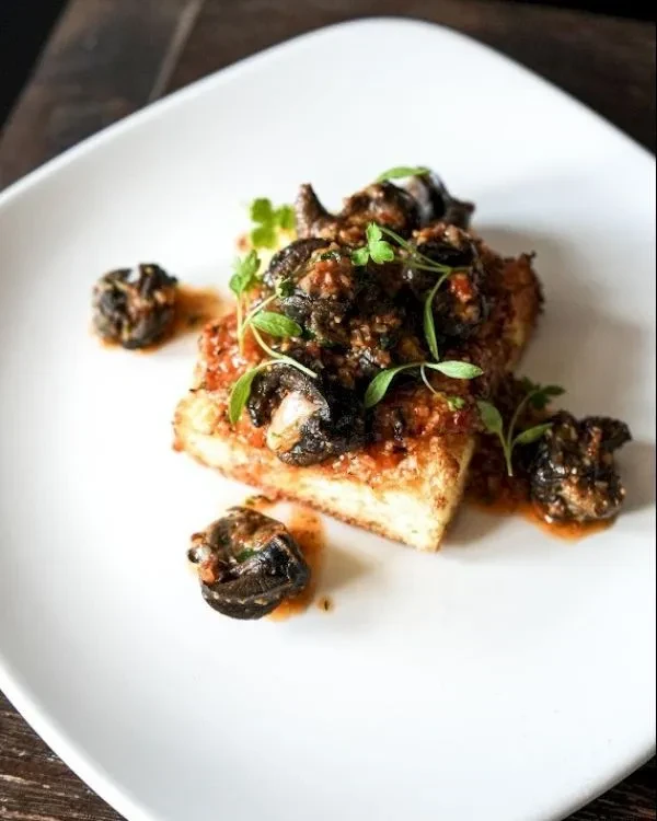 White plate with rectangular food, dark savory topping, and herbs.